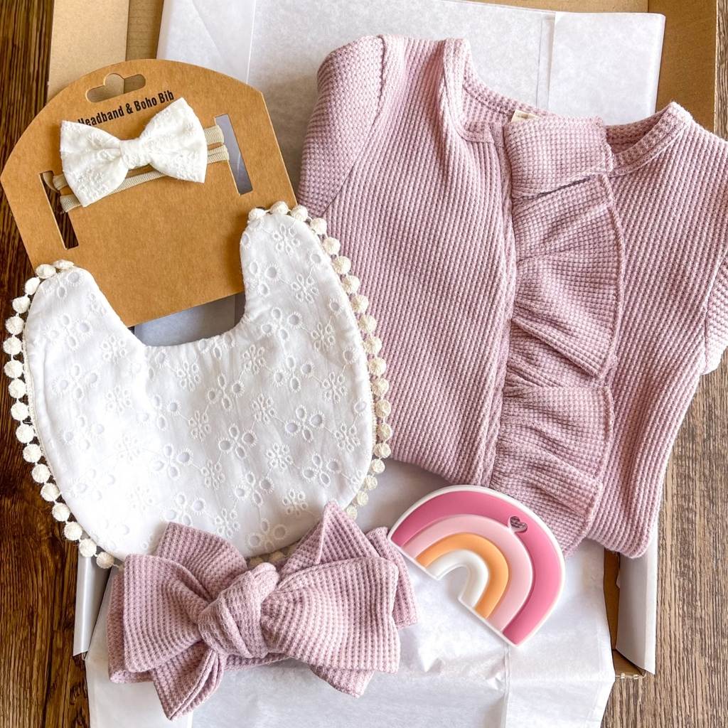 Light Purple 5 piece baby girl gift box with white lace bib and matching headband, thermal knit ruffled jumpsuit with matching headband and a pink silicone rainbow toy.